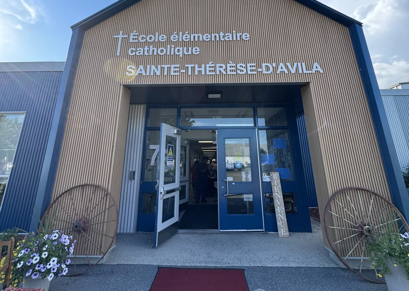 Vue de l'entrée principale de l'École élémentaire catholique Sainte-Thérèse-d'Avila. La façade est revêtue de lattes de bois verticales avec le nom de l'école affiché en grandes lettres blanches. Un tapis rouge mène aux portes d'entrée ouvertes, encadrées de deux grandes roues de chariot décoratives et de bacs de fleurs.