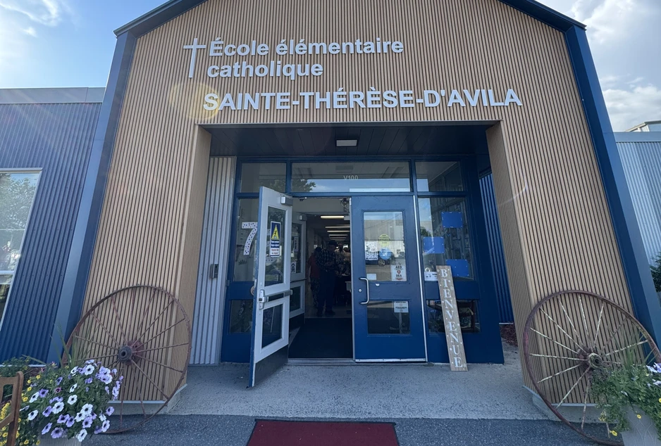 Vue de l'entrée principale de l'École élémentaire catholique Sainte-Thérèse-d'Avila. La façade est revêtue de lattes de bois verticales avec le nom de l'école affiché en grandes lettres blanches. Un tapis rouge mène aux portes d'entrée ouvertes, encadrées de deux grandes roues de chariot décoratives et de bacs de fleurs.
